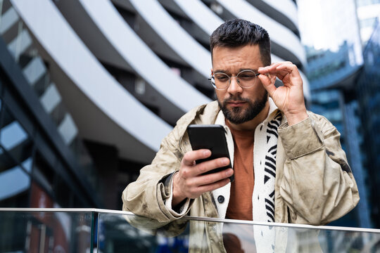 Young professional in a modern business district checks his phone—smiling at good news and managing messages, payments, maps, and schedules on the go while commuting between meetings across the city