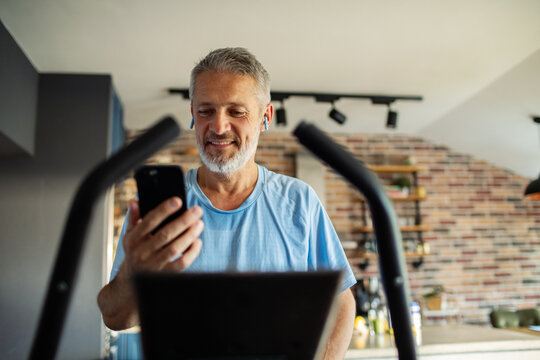 Mature man smiling on elliptical with smartphone at home
