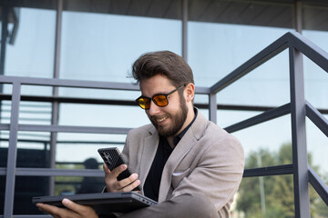 A man with a beard and sunglasses is sitting outdoors, looking at his smartphone