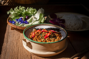 A plate of traditional Southern Thai Khua Kling stir-fried minced pork served in a rustic bowl, topped with red chili and sliced kaffir lime leaves. Fresh vegetables such as lettuce, eggplants, and bu