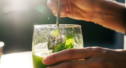 Person stirring a green smoothie in a glass with a spoon on a sunny day