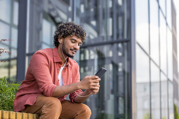 A young Indian man in casual clothes sits with headphones on a bench near a building and uses his phone with a smile