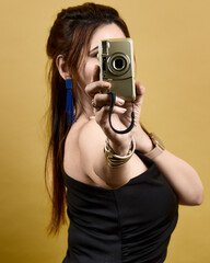 Studio portrait of a stylish woman holding a camera toward the viewer, set against a golden backdrop. Represents empowerment, creativity, and self-confidence in a modern fashion style.