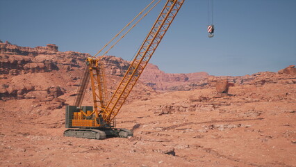 A towering crane stands in a vast desert with rocky formations and red sand. Its arm stretches high as it prepares to lift a heavy load against a backdrop of distant mountains.