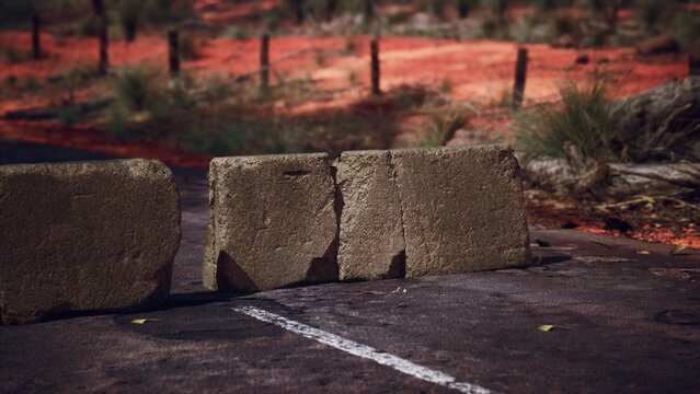 A lonely road in the Australian outback is blocked by a row of large stones. The bright red earth and sparse vegetation showcase the rugged beauty of this remote landscape under the midday sun.