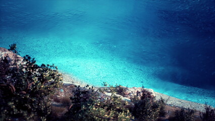 Tranquil shoreline features shimmering azure water with gentle waves lapping the pebbled beach, surrounded by lush greenery under a clear blue sky, perfect for relaxation.
