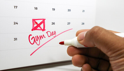 A hand marking Gym Day and crossing out the 25th on a calendar with a red marker.