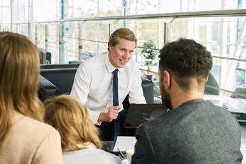 Caucasian young adult man smiling and explaining contract details to multiethnic family including Caucasian woman, Black man, and child while sitting at car dealership table