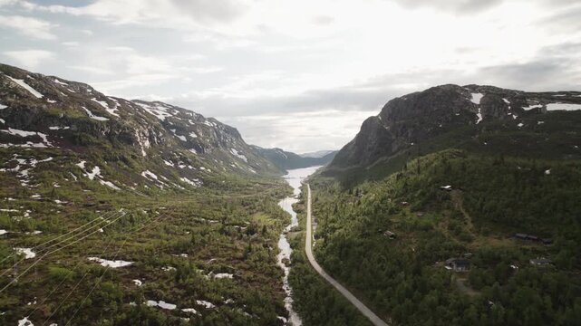 Aerial view of a winding road that cuts through a lush valley with mountains and a lake in the distance, Dalen, Telemark, Norway.