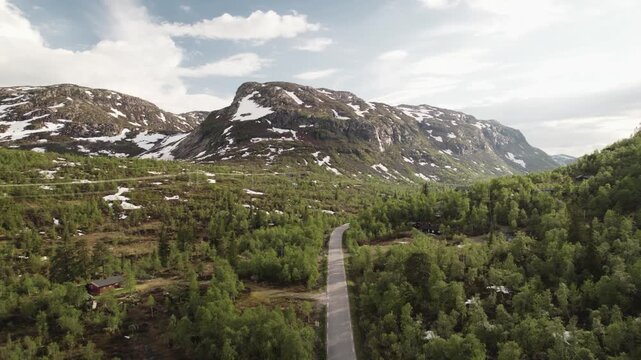 Aerial view of a winding road through a green landscape, contrasting with snow-capped mountains in the distance, Dalen, Telemark, Norway.