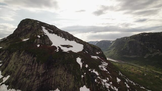 Aerial view of a serene lake nestled between rugged mountains, where patches of snow meet lush greenery under a cloudy sky, Dalen, Telemark, Norway.