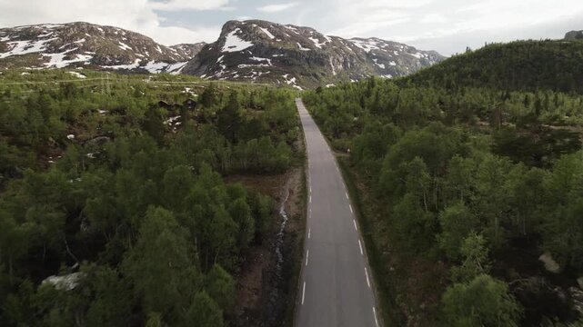Aerial view of a long grey road cutting through lush green trees, leading towards mountains with patches of snow, Dalen, Telemark, Norway.
