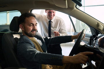 Middle aged Caucasian man sitting in driver seat of car smiling and interacting with dashboard controls while middle aged Caucasian man