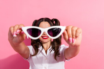 Bright young woman in white shirt holding pink sunglass toward camera against pink background smiling and playful