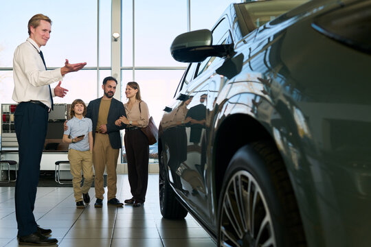 Caucasian male car salesman presenting vehicle to Middle aged Caucasian man, Middle aged Caucasian woman, and child boy standing together inside modern car dealership showroom