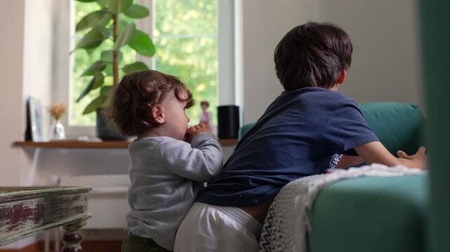 Toddler reaching toward older sibling leaning on couch, seeking attention with playful persistence and affection, expressing curiosity and connection in warm family home environment