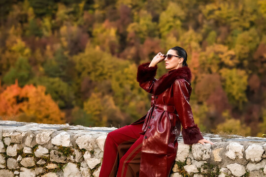 Stylish woman in a burgundy leather coat sitting on an old stone wall, adjusting her sunglasses, with a colorful autumn forest behind her. Elegant outdoor fashion portrait in warm natural light.