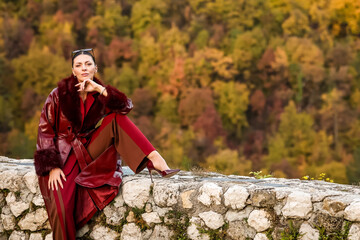 Elegant woman in a burgundy leather coat sitting on a stone wall, posing confidently against a colorful autumn forest. Stylish fall fashion portrait in warm natural light.