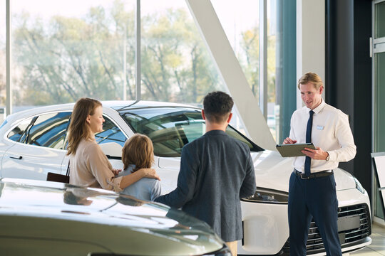 Caucasian male car salesman standing with clipboard assisting Caucasian middle aged man, Caucasian woman, and Caucasian child inside car dealership showroom near new vehicles