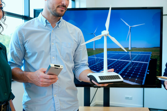 Caucasian young adult man holding wind turbine model with solar panel, standing in office with colleagues, presenting renewable energy concept in front of large display screen