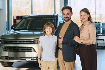 Caucasian middle aged man holding car key standing with smiling Caucasian woman and Caucasian boy in car dealership showroom, family posing together in front of new vehicle
