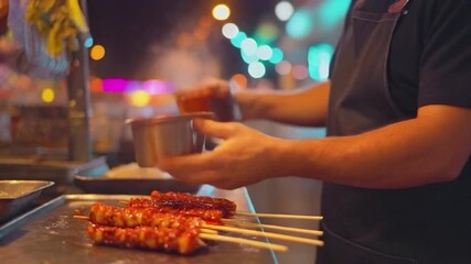 Street food vendor basting skewered meat on a hot griddle at night