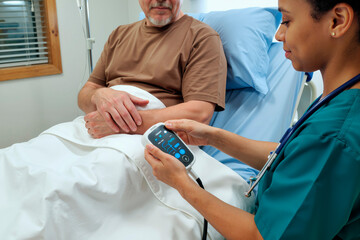 Middle aged Caucasian man lying in hospital bed receiving care from young adult Black woman nurse checking vital signs with digital medical device in clinical setting