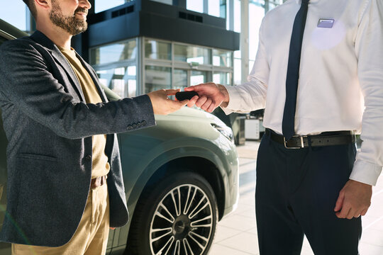 Caucasian middle aged man receiving car keys from Caucasian young adult man in dealership showroom, both standing near vehicle, business transaction taking place, hands visible