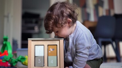 Toddler sitting on floor beside wooden activity cube, deeply focused while exploring numbers and shapes during playful early learning moment at home