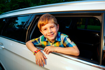 Portrait of Caucasian boy child smiling while leaning out of car window during sunny day, showing colorful beaded bracelets on wrist, looking directly at camera outdoors
