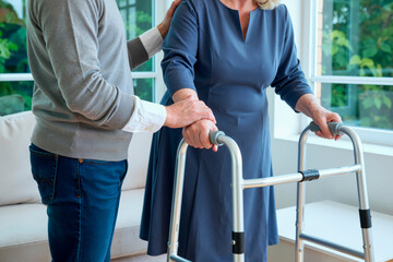 Senior Caucasian woman using walker for support while middle aged Caucasian man assisting her indoors, both standing near window with greenery visible outside, focusing on mobility aid