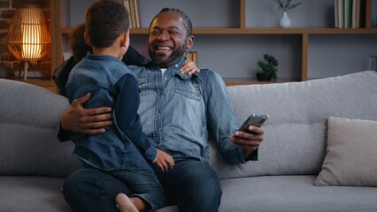 Happy African American family daddy using phone sons jumping running surprise dad cuddling laughing together with mobile smartphone two boys kids children at home enjoying relationship father's day