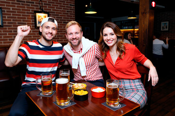 Three Caucasian young adults sitting at table smiling and posing for camera, enjoying beer and snacks in pub, two men and one woman showing cheerful expressions together