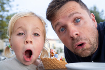 Dad and Little Girl Sitting in a Cafe, Eating Ice Cream and Looking Surprised at the Camera
