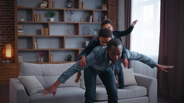 Carefree African American family playing piggyback pretending airplane at home joyful father dad riding on back little son play plane flying pretend with two kids boys sons children having fun joy