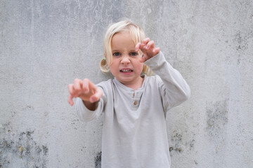 Little 4 Year Old Blonde Girl Standing by a Wall, Pretending to Be Angry and Showing Claws