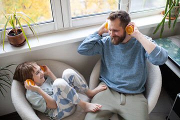 Bearded father and son playing with oranges in bright kitchen. Man and boy holding two orange halves near head like ears. Family life, homemade juice, vitamin C, home lifestyle.