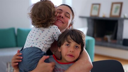 Mother holding toddler and older child together in warm family embrace, smiling softly while both children cuddle close in affectionate moment at home