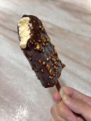 Hand holding a chocolate coated vanilla ice cream bar with nuts and a bite taken.