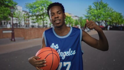 Man holds basketball on city street in blue jersey amid urban trees and buildings; confidence pride focus drive. - Powered by Adobe