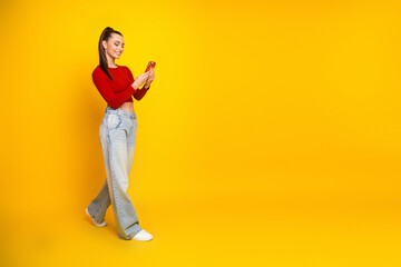 Young woman in red top and jeans smiles while using a smartphone against a bright yellow background in a casual fashion lifestyle scene © deagreez