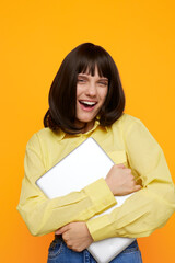 Fototapeta premium A cheerful young woman in a yellow shirt hugs a silver laptop against a bright orange backdrop, conveying enthusiasm, study motivation, and modern technology for creative projects.