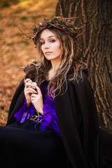 A young woman stands in a golden autumn forest, wearing a black medieval dress with purple accents. She wears a crown of twigs and berries, and a dark hooded cloak. This fantasy portrait  