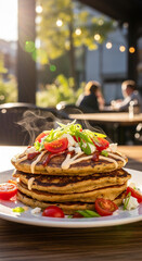 Savory stack of fluffy pancakes topped with fresh tomatoes, feta cheese, and green onions, served outdoors with blurred diners in background