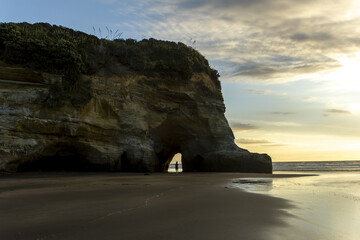 View of a silhouette standing within a naturally carved archway in a coastal cliff face, where the beach reflects the sky, Tongaporutu, Taranaki Region, New Zealand.
