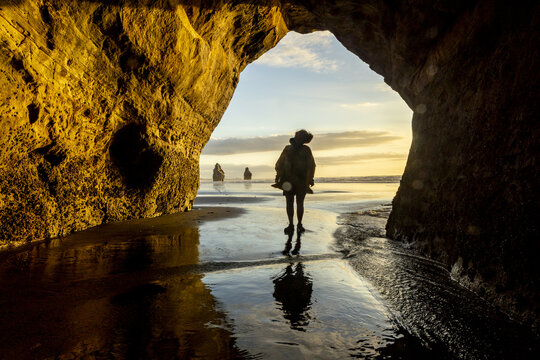 View of a person silhouetted against the ocean framed by a golden cave entrance where the wet sand reflects the sky, Tongaporutu, Taranaki Region, New Zealand.