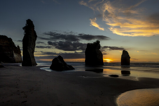 View of monolithic sea stacks rise majestically from the dark, wet sands under a sky ablaze with the dying embers of sunset, Tongaporutu, Taranaki Region, New Zealand.