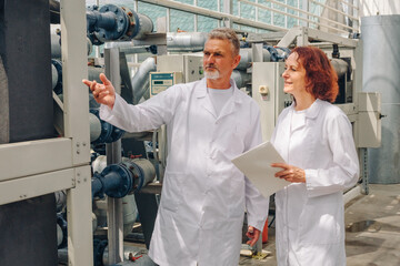 Senior engineers inspecting heavy machinery controls, assessing reliability and lifecycle needs, pointing at components and noting service actions on clipboard in industrial maintenance area