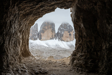 View of the Tre Cime di Lavaredo peaks are framed by a rugged cave entrance, with snow-dusted slopes under a cloudy sky, Auronzo di Cadore, Province of Belluno, Italy.
