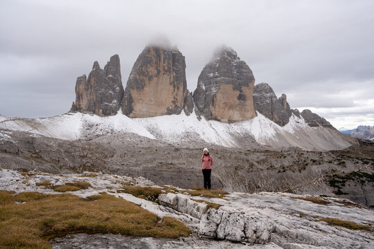 View of a person standing amidst the rugged, snow-dusted landscape, with towering Tre Cime peaks veiled in mist under a clouded sky, Auronzo di Cadore, Province of Belluno, Italy.